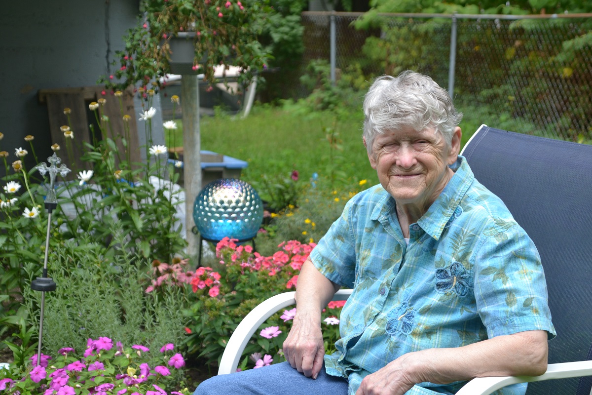 Vanessa Munsey sits in her garden. Behind her are pink and white flowers and a garden globe.